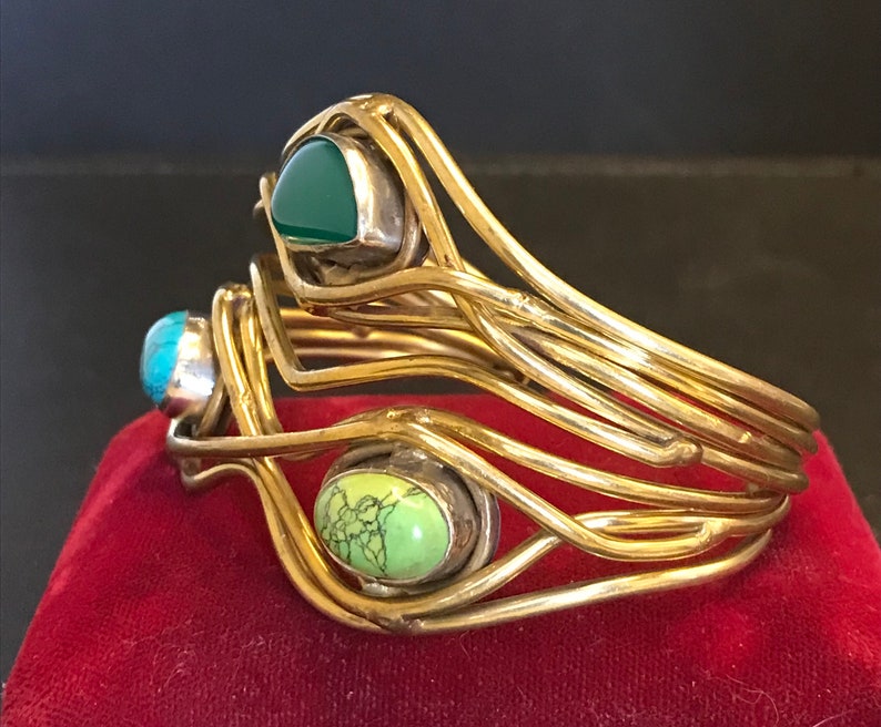 May include: A gold-toned bangle bracelet with three different colored stones. The stones are green, turquoise, and a light green color. The bracelet is made of multiple strands of wire that are woven together.