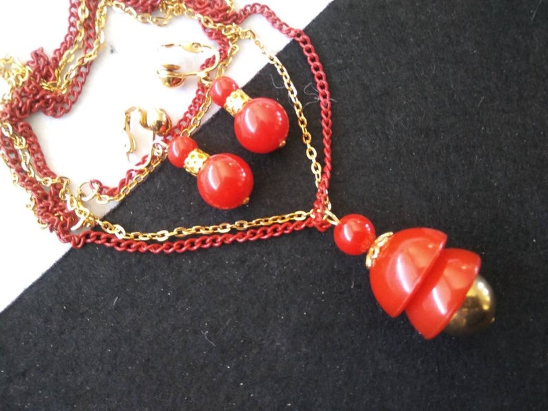 May include: A red and gold necklace and earrings set. The necklace has three chains, two gold and one red. The pendant is a red, layered, teardrop shape with a gold accent. The earrings are clip-on and feature two red beads.