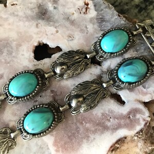 Op de afbeelding: Close-up van een vintage zilverkleurige armband met ovale turquoise stenen. De armband heeft een gedetailleerd blad- en kralenontwerp tussen de stenen, tegen een lichtgekleurde, getextureerde achtergrond.