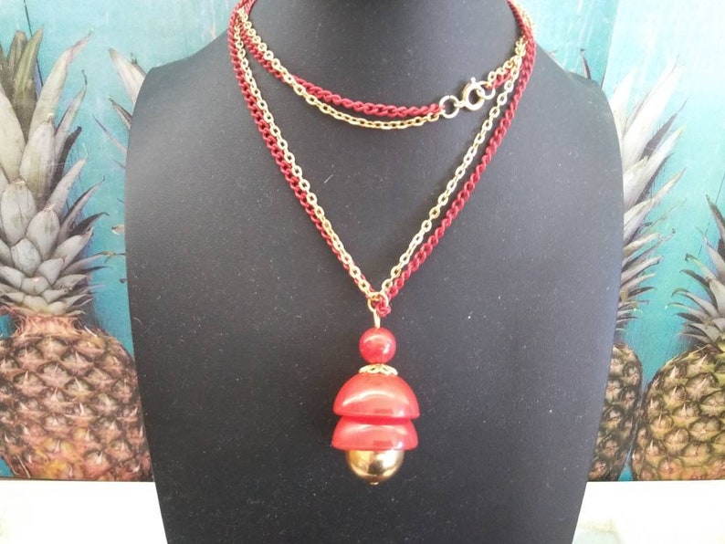 May include: A layered necklace with a red and gold pendant. The pendant is made of three stacked red circles with a gold ball at the bottom.