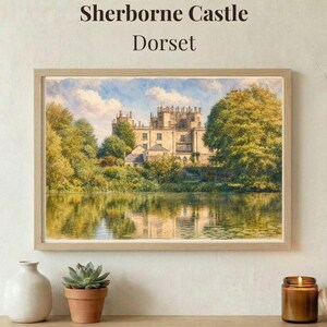 May include: A framed landscape painting titled Sherborne Castle Dorset depicting a stone castle surrounded by lush green trees and reflected in a calm body of water under a blue sky with white clouds. A white vase and a succulent are nearby.
