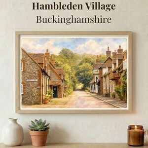 Op de afbeelding: Een ingelijste aquarelprint getiteld Hambleden Village Buckinghamshire met een stenen huisjesstraat, een zandpad, weelderige groene bomen en een blauwe lucht met wolken. Het kunstwerk is ingelijst in een lichtgekleurde houten lijst.