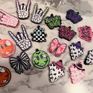 May include: A collection of colorful silicone charms featuring skeleton hands, ghosts, smiley faces, bows, and cow print patterns. Several charms display the text Spooky Babe. These decorative items include various shapes like circles, bows, and hands.
