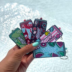May include: A collection of five rectangular keychains shaped like beverage cans, each featuring the brand name Alani in white script. The designs include green with lime slices, red with cherries, dark leopard print, pink stripes, and blue with watermelon.