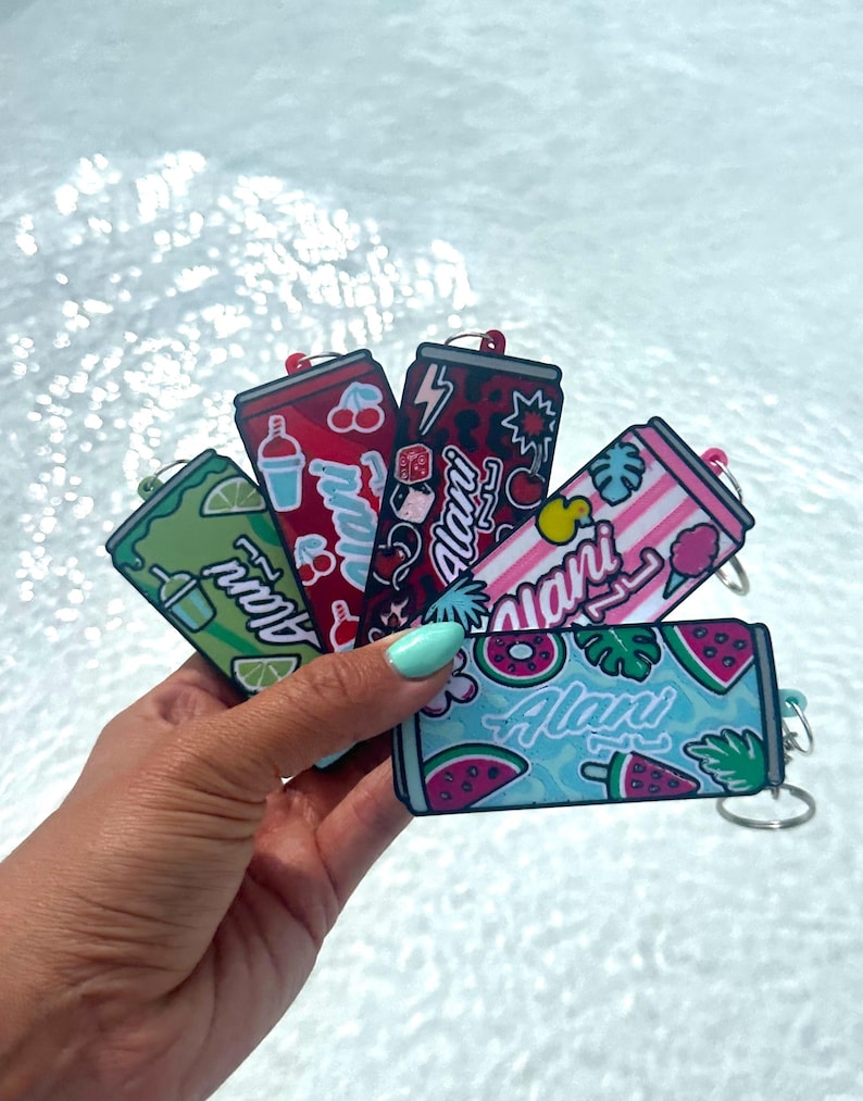 May include: A collection of five rectangular keychains shaped like beverage cans, each featuring the brand name Alani in white script. The designs include green with lime slices, red with cherries, dark leopard print, pink stripes, and blue with watermelon.