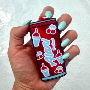 May include: A red rectangular keychain shaped like a beverage can featuring the word Alani in light blue script, surrounded by small illustrations of light blue slushy drinks and pairs of red cherries, finished with a black border and metal ring.