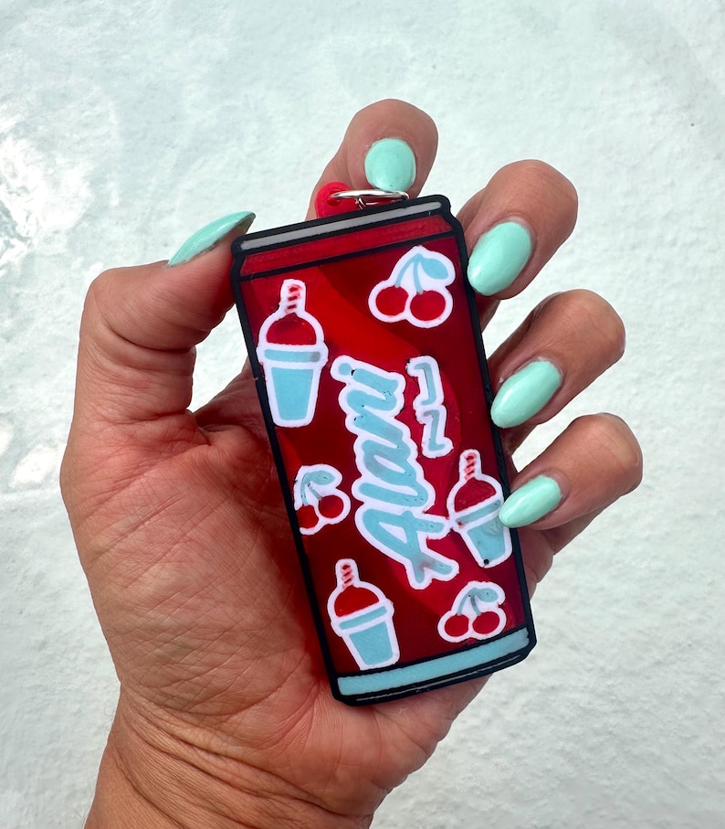May include: A red rectangular keychain shaped like a beverage can featuring the word Alani in light blue script, surrounded by small illustrations of light blue slushy drinks and pairs of red cherries, finished with a black border and metal ring.