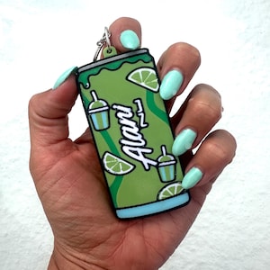 May include: A flat, rectangular keychain shaped like a beverage can in shades of green and light blue. The design features the word Alani in white script, along with illustrations of lime wedges and small drink cups with straws on a green background.
