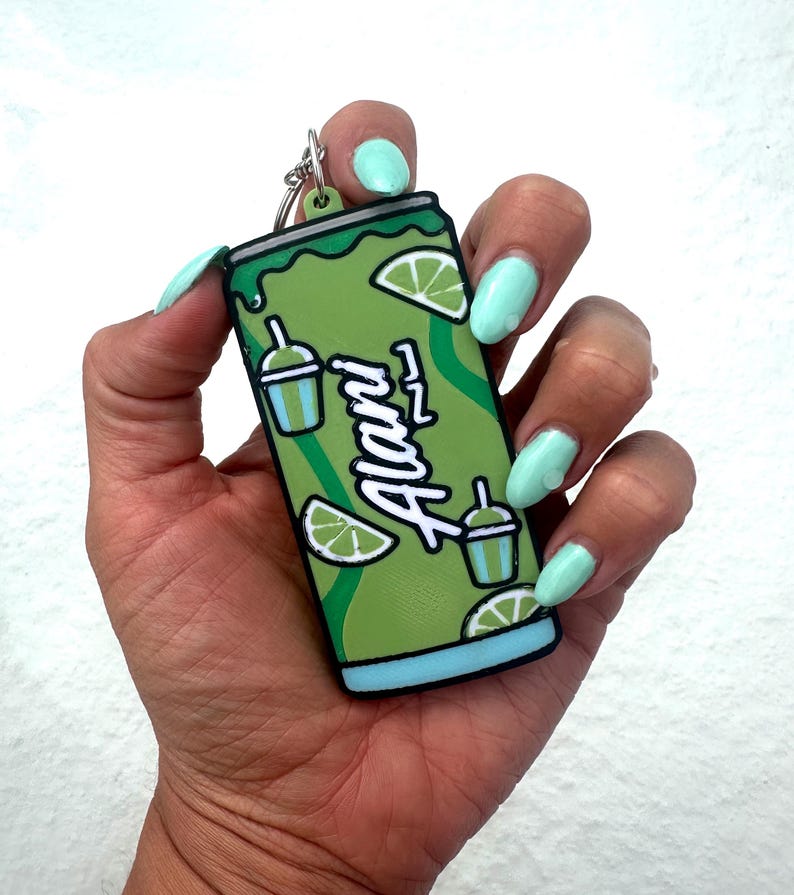 May include: A flat, rectangular keychain shaped like a beverage can in shades of green and light blue. The design features the word Alani in white script, along with illustrations of lime wedges and small drink cups with straws on a green background.
