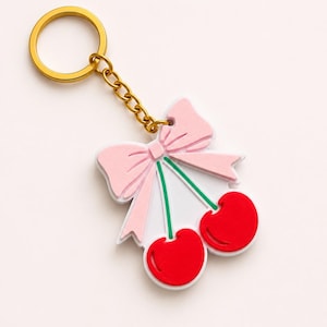 May include: A keychain featuring a gold-toned metal ring and chain attached to a flat, layered charm. The charm depicts two bright red cherries with green stems, topped with a light pink bow. The entire cherry and bow design has a white border.