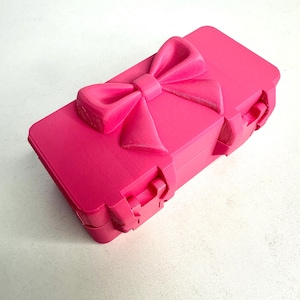 Puede incluir: Una caja rectangular rosa brillante con un gran lazo en la tapa. La caja tiene un cierre de cierre y está hecha de un material liso. El lazo es un elemento de diseño tridimensional prominente.