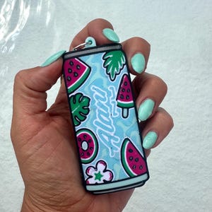 May include: A rectangular keychain shaped like a beverage can with a light blue background featuring a pattern of watermelon slices, green leaves, and flowers. The word Alani is written in white script across the center of the can design.