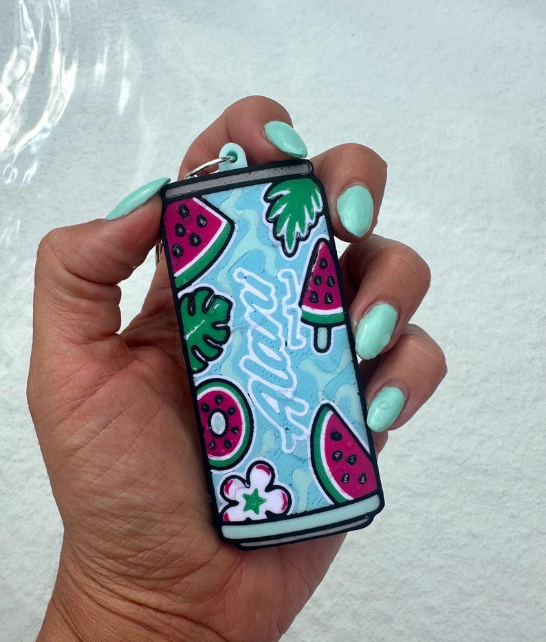 May include: A rectangular keychain shaped like a beverage can with a light blue background featuring a pattern of watermelon slices, green leaves, and flowers. The word Alani is written in white script across the center of the can design.