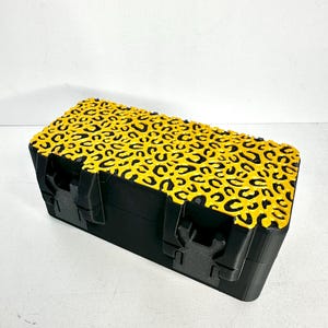 May include: A black rectangular box with a yellow lid featuring a black leopard print design. The box has two black clasps on the front. The box is sitting on a white surface.