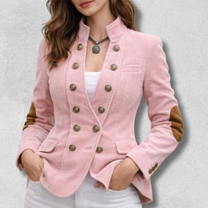 Puede incluir: Blazer de pana rosa claro con diseño de doble botonadura. La chaqueta tiene botones dorados, cuello alto y coderas marrones. La modelo lleva un top blanco y pantalones blancos.