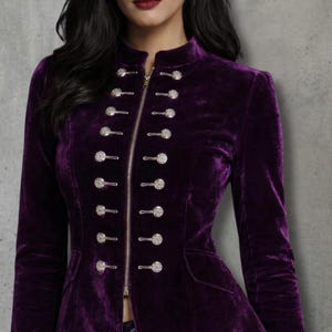 Puede incluir: Chaqueta de terciopelo morado oscuro con cuello alto y botones plateados decorativos. La chaqueta tiene una silueta entallada, cierre de cremallera y mangas largas. La tela parece suave y lujosa. Diseño elegante y con estilo.