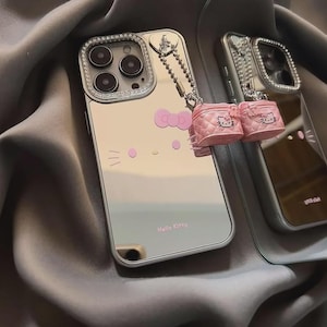 May include: Silver phone case with a Hello Kitty design. The case has a pink bow, pink Hello Kitty face, and the words "Hello Kitty". A small pink purse charm hangs from the case. The camera lens is surrounded by a ring of clear crystals.