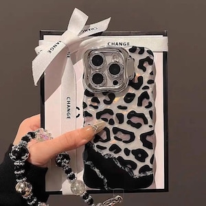 May include: A phone case featuring a leopard print pattern in black, white, and pink. The case has a clear camera lens protector with a silver frame. It is presented with a white ribbon printed with the word "CHANGE".