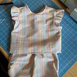 May include: A pastel striped two-piece outfit, comprising a top with ruffled sleeves and matching shorts. The fabric features vertical stripes in pink, blue, yellow, and green. The outfit is laid out on a blue cutting mat.