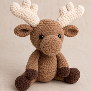 May include: A crocheted stuffed animal in the shape of a reindeer. The body is medium brown with dark brown feet, while the antlers are cream-coloured. It features two small black plastic eyes and a rounded snout, crafted from soft yarn.