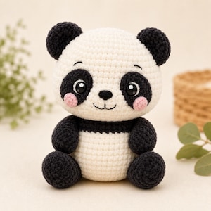 May include: A crocheted panda stuffed animal featuring a round white head with black ears, black eye patches, and small pink cheeks. The body is white with black arms and legs, crafted from yarn with a visible textured stitch pattern throughout.