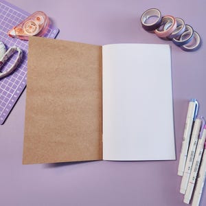 May include: An open notebook with brown and white pages, surrounded by stationery items. The items include washi tape rolls in various colors, pens, a cutting mat, and a tape runner, all on a purple surface.