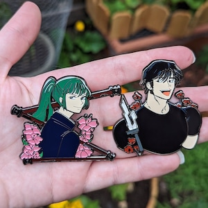 Może przedstawiać: Dwie emaliowane przypinki przedstawiające postacie w stylu anime. Jedna przypinka przedstawia postać z zielonymi włosami i różowymi kwiatami, druga postać z czarnymi włosami i czerwonymi kwiatami. Obie przypinki mają czarny kontur.
