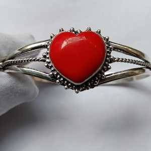 May include: A silver-toned cuff bracelet featuring a vibrant red heart-shaped centerpiece. The heart is surrounded by a decorative border, and the bracelet has a twisted rope design on the sides. The jewelry is displayed on a white background.