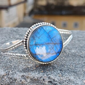 May include: A silver cuff bracelet featuring a large, round, iridescent blue labradorite gemstone at the center. The metal band has a twisted rope texture and a beaded border surrounding the stone, creating a detailed and polished appearance.