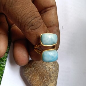 May include: A gold-colored ring featuring two rectangular, light blue gemstones. The gemstones are set in a gold bezel setting. The ring is held up by a person's fingers.