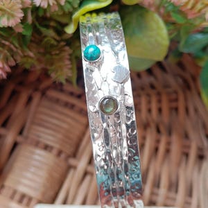 May include: A silver bracelet with a textured, hammered finish. It is adorned with a turquoise stone, a small heart-shaped detail, and a green stone. The bracelet is set against a background of foliage and a wicker basket.