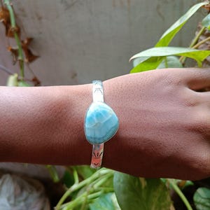 Pode incluir: Uma pulseira de punho de prata com uma grande pedra de larimar azul claro. A pedra tem uma forma orgânica única e uma aparência marmoreada. A pulseira tem padrões decorativos gravados na banda.