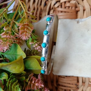 May include: A silver cuff bracelet featuring five turquoise gemstones. The bracelet is displayed against a neutral backdrop, with green and pink foliage in the foreground. The design is simple and elegant, suitable for everyday wear.