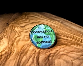 May include: A round pin with a blue and green world map design. The pin has the words "Compassion has no borders" in white and green text. The pin is on a wooden surface.