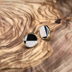 Puede incluir: Dos pendientes de botón redondos con borde dorado. Cada pendiente presenta un diseño geométrico en blanco y negro. Los pendientes se muestran sobre una superficie de madera con un patrón de vetas natural.