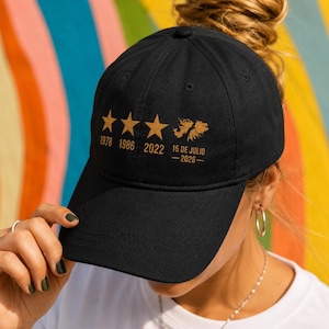 May include: A black baseball cap featuring three gold stars above the years 1978, 1986, and 2022. To the right of the stars is a gold map outline, with the text 15 DE JULIO - 2026 printed below it in a gold sans-serif font.