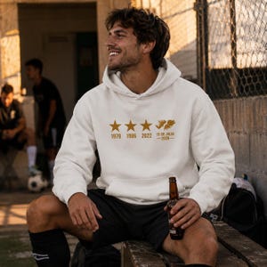 May include: A white hooded sweatshirt featuring a gold graphic print on the chest. The design includes three stars above the years 1978, 1986, and 2022, followed by a map silhouette and the text 15 DE JULIO 2026. The hoodie has long sleeves.