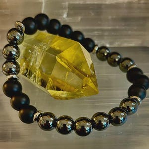 May include: A beaded bracelet featuring alternating matte black and metallic gray beads. The bracelet is accented with silver-toned spacers. A large, faceted yellow crystal is in the background, adding a contrast of color and texture. The bracelet is likely a piece of jewelry.