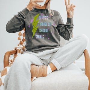 Könnte beinhalten: Ein dunkelgraues Langarmshirt mit dem Text "Dancing Through The Lightning Strikes" und einem neongrünen Blitz-Grafik. Das Shirt wird mit grauer Jogginghose und einer weißen Mütze kombiniert. Die Person sitzt in einem Holzstuhl.