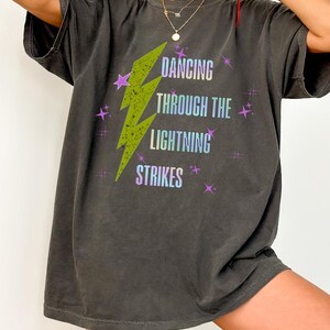 Könnte beinhalten: Dunkelgraues T-Shirt mit dem Text "DANCING THROUGH THE LIGHTNING STRIKES" in einer Farbverlaufs-Schriftart. Ein grüner Blitz ist mittig auf dem Shirt, mit lila Sternakzenten. Das Shirt hat eine lockere Passform.
