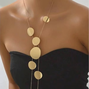 Puede incluir: Un collar dorado con una serie de colgantes planos en forma de disco de forma irregular. Los colgantes están suspendidos de una delicada cadena, creando un efecto en capas. El collar se lleva sobre un top negro sin tirantes.