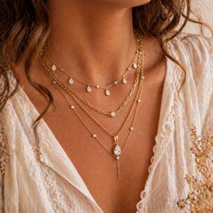 Op de afbeelding: Een gelaagde gouden ketting set met meerdere kettingen met verschillende ontwerpen. Sommige kettingen hebben kleine, heldere edelstenen, andere hebben een druppelvormige hanger. De kettingen worden gedragen door een persoon met een crèmekleurige top.