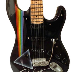 Puede incluir: Miniatura de guitarra eléctrica negra con una franja de arcoíris y el logotipo de Pink Floyd. La guitarra tiene mástil y diapasón de madera, con herrajes plateados y una palanca de trémolo. Un artículo decorativo para los amantes de la música.