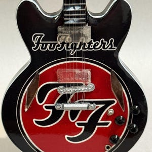 May include: Miniature black electric guitar with a red circular design featuring the band's logo. The guitar has the band name "Foo Fighters" in white script. The guitar has a white trim and silver hardware.