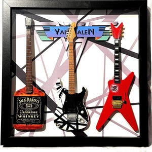 May include: A framed artwork featuring three miniature electric guitars. One guitar is shaped like a Jack Daniel's whiskey bottle, another has a black and white design, and the third is red. The Van Halen logo is above the guitars.