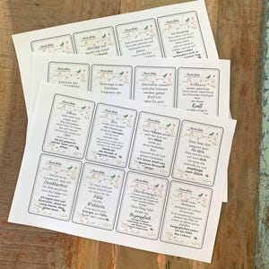 May include: Several printed cards with floral borders and text in German are stacked on a wooden surface. The cards feature inspirational quotes and affirmations. The cards are rectangular with rounded corners and a white background.