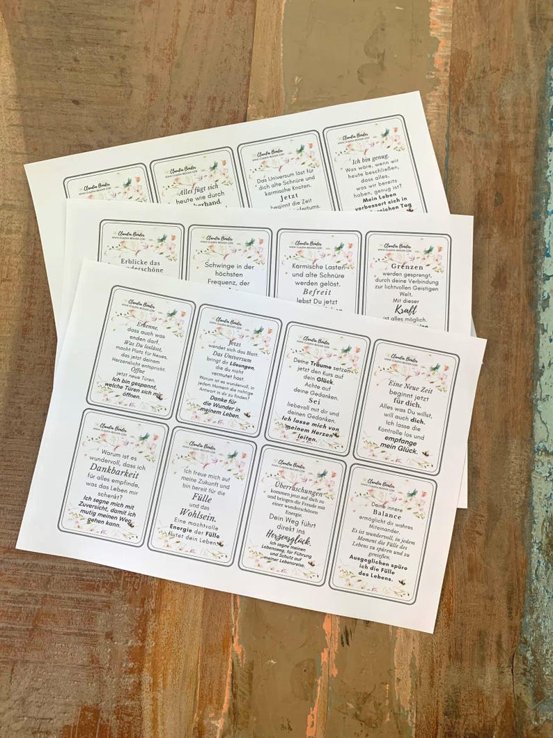 May include: Several printed cards with floral borders and text in German are stacked on a wooden surface. The cards feature inspirational quotes and affirmations. The cards are rectangular with rounded corners and a white background.
