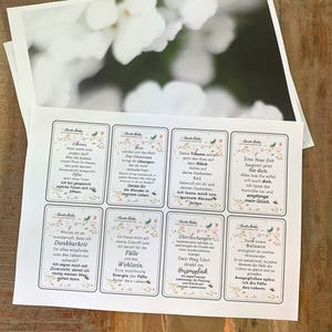 May include: A collection of printed cards with inspirational quotes and floral designs, alongside a blurred photograph of white flowers. The cards feature phrases in German, with a soft, neutral color palette. The cards are arranged on a wooden surface.