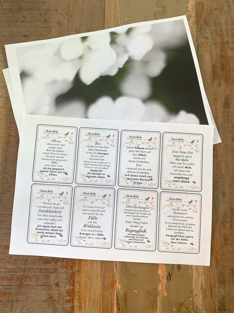 May include: A collection of printed cards with inspirational quotes and floral designs, alongside a blurred photograph of white flowers. The cards feature phrases in German, with a soft, neutral color palette. The cards are arranged on a wooden surface.