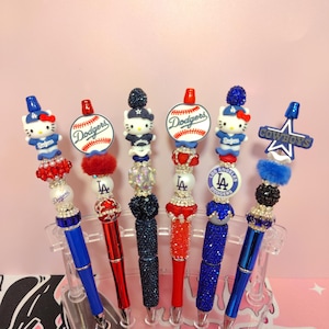 May include: Six decorative pens featuring sports team themes, including Hello Kitty figures, baseballs, and team logos. The pens are adorned with colorful beads, rhinestones, and glittery accents in blue, red, and white, displayed in a clear plastic stand.